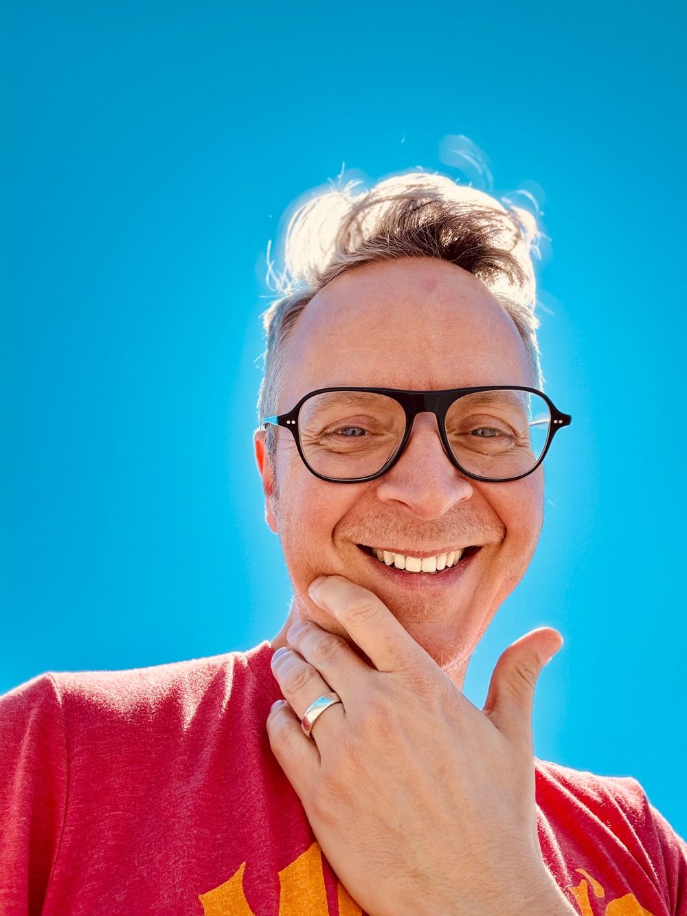 Marcus Greatheart smiling outdoors against a blue sky, wearing glasses and a red t-shirt
